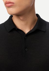 Gros plan sur une personne portant un pull noir à col avec deux boutons visibles, montrant le cou et la partie inférieure du visage.