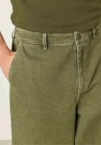 Olivově zelené bavlněné kalhoty s volnějším střihem, vybavené zapínáním na jeden knoflík, předními kapsami a kontrastními švy.