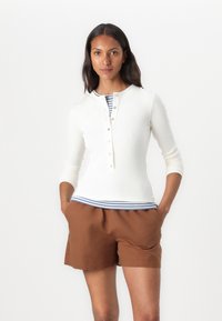 Hvit ribbet langermet topp med knappestolpe lagt over en blåstripete skjorte, parret med brune shorts. Uformell, tettsittende stil.
