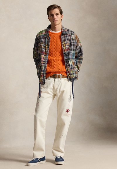 Ung mann som har på seg en flerfarget lappejakke, en skarp oransje kabelstrikket genser, kremfargede bukser med broderidetaljer, og blå joggesko.