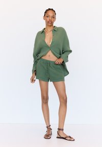Green woven cotton short set featuring a cropped button-up shirt and matching shorts. Model wears brown strappy sandals and a gold necklace.