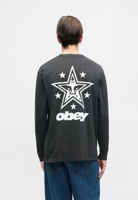 Camisa de manga longa cinza-escuro com um grande gráfico de estrela branca, com cinco estrelas e o texto "obey" nas costas. Material de algodão, corte relaxado.