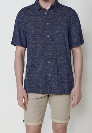 Homme portant une chemise bleu marine à manches courtes avec un motif subtil et un short beige retroussé, debout devant un fond uni.
