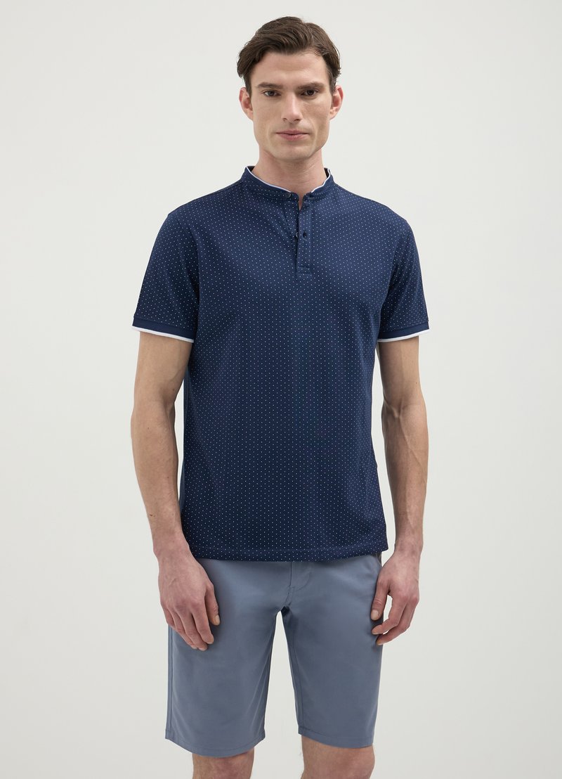 Polo shirt bleu marine à manches courtes avec col mandarin, motif à pois discret et accents blancs sur les manches. Porté avec un short gris clair.
