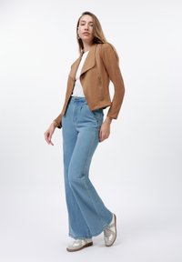 Femme debout dans une veste en daim marron, un haut blanc, un jean flare bleu clair taille haute et des baskets blanches devant un fond blanc.