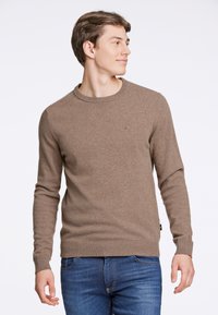 Brun stickad tröja med rund halsringning, långa ärmar och en subtil logotypdetalj på bröstet. Matchad med blå jeans.