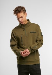 Felpa a collo alto color verde oliva con zip a metà davanti, tasca sul petto e patch in velcro sulle maniche. Tessuto morbido e testurizzato.