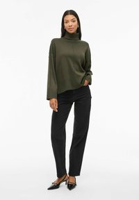 Un pull col roulé oversize vert foncé, avec des poignets côtelés, associé à un jean noir à jambe droite et des chaussures à talons noires.