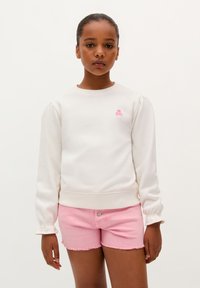 Weißes Sweatshirt mit Puffärmeln, gerippten Bündchen und kleinem pinken Totenkopf-Logo; kombiniert mit pinken Jeansshorts mit ausgefransten Säumen.