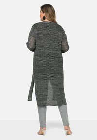 Dlouhý otevřený pletený kardigan v černé a šedé barvě, s volným střihem, bočními rozparky a lehkou texturovanou tkaninou, kombinovaný se šedými kalhotami.