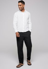 Camisa blanca de lino de manga larga, abotonada, con textura ligera; combinada con pantalones de lino gris oscuro y chanclas negras.