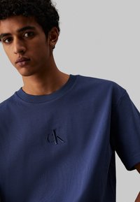 Camiseta de algodón azul marino con cuello redondo y mangas cortas, que presenta un logo bordado de 'cK' en la parte izquierda del pecho.