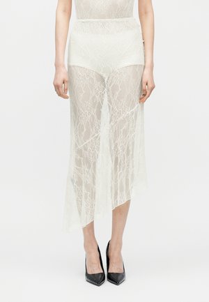 Femme portant une jupe asymétrique en dentelle blanche par-dessus un short blanc taille haute et des talons noirs à bouts pointus, debout devant un fond uni.