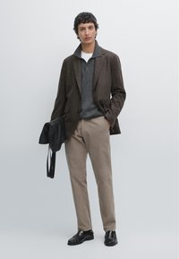 Hnědý blazer s pruhy přes šedý svetr, doplněný krémovými kalhotami a černými mokasíny. Modelka drží černou tašku se šňůrkami.