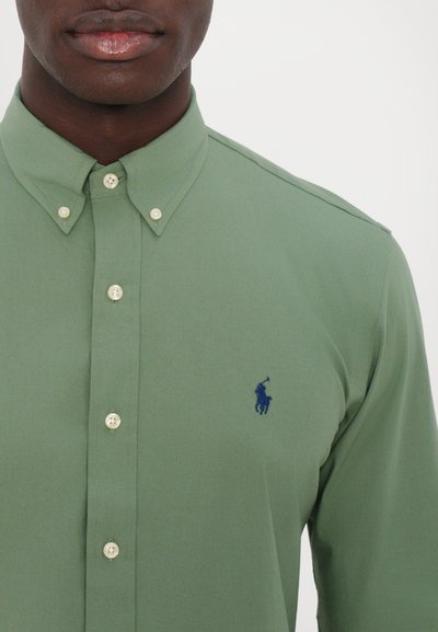 Chemise verte à boutons avec col, ornée d'un petit logo bleu sur la poitrine gauche et de boutons de couleur claire. Texte de tissu lisse.
