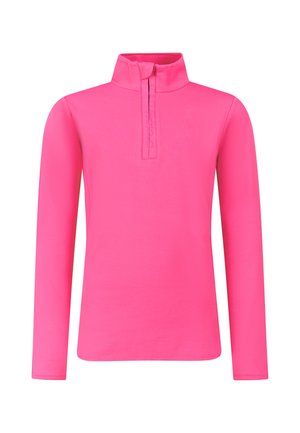 Jonge vrouw staat met ontspannen handen, draagt een felroze top met lange mouwen, donkerpaarse broek en zwarte schoenen op een lichte achtergrond.