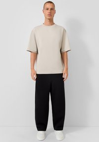 Beige Übergröße T-Shirt mit kurzen Ärmeln, kombiniert mit lockeren schwarzen Hosen und weißen Sneakers, minimalistisches Design, glatte Textur.