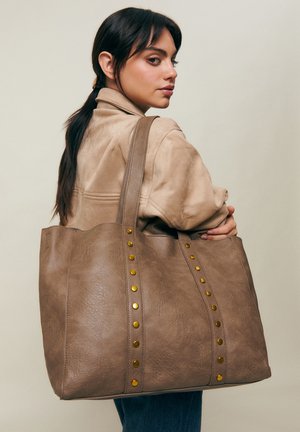 Žena s tmavými vlasmi stiahnutými do copu, oblečená v béžovom saku a modrých džínsach, nesie veľkú hnedú koženú tašku s pozlátenými cvokmi na ramene.