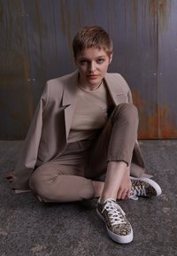 Beige blazer over a ribbed top, matching beige trousers. Sneakers with a snake pattern and white laces. Texture contrasts with the background.