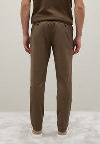 Bruine slim-fit broek met een gladde textuur, voorzien van twee achterzakken en zwarte knoopaccenten, gecombineerd met witte schoenen.