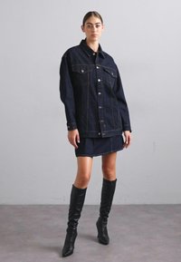 Jachete de ganga escura com dois bolsos no peito, botões prateados e costuras contrastantes, combinada com uma saia mini de ganga e botas pretas até ao joelho.