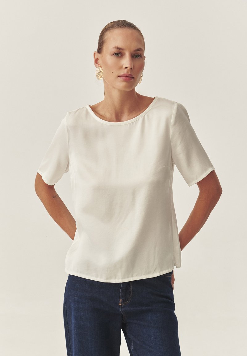 Blusa blanca de manga corta con cuello redondo, textura suave y ligero brillo, combinada con unos vaqueros de mezclilla azul oscuro.