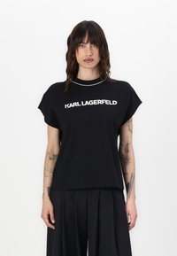 Melnā, īsa piedurkņu T-kreklā ar apaļu kakla izgriezumu, uz kura priekšpuses ir uzdrukāts uzbolds balts teksts "KARL LAGERFELD". Mīksts audums, brīvs griezums.
