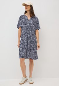 Abito blu navy con motivo a foglia bianca, maniche corte, scollo a V e vita elasticizzata. Il modello indossa un cappello beige e sneakers bianche con suola beige.