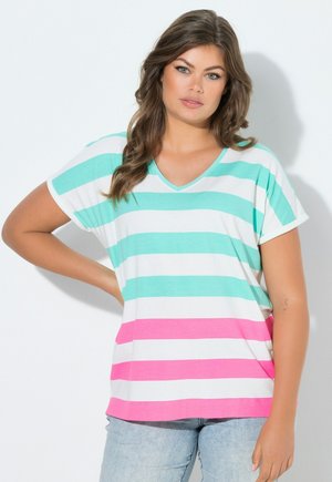 Donna con capelli lunghi e mossi che indossa una maglietta a V con strisce orizzontali bianche, turchesi e rosa, posando con una mano sul fianco su sfondo bianco.