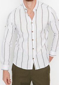 Valkoinen nappipaita pystysuorilla harmailla raidoilla, jossa on kaulus, pitkät hihat ja ruskeat nappikoristeet. Rakennekangas lisää visuaalista kiinnostavuutta.