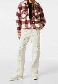 Rutig jacka i bordeaux och kräm med två bröstfickor; i kombination med vita cargobyxor som har flera fickor och dragkedjor.