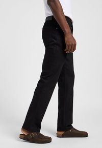 Seitliche Ansicht einer Person, die schwarze Jeans, ein weißes Hemd und braune Slipper mit Schnalle trägt, stehend vor einem einfarbigen Hintergrund.