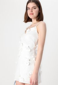 Vestido mini blanco con escote en V, tirantes finos y apliques florales en 3D. Tejido suave con un diseño ajustado y acentos de flores texturizadas.