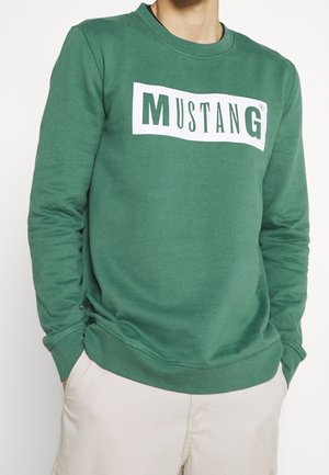 Mann trägt ein grünes Sweatshirt mit weißem rechteckigem Aufnäher, auf dem "MUSTANG" in grünen Buchstaben steht, kombiniert mit hellbeigen Hosen.