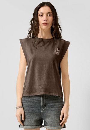 Femme aux longs cheveux foncés portant un haut marron sans manches avec des épaulettes et un short en jean retroussé sur un fond uni.