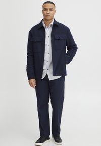 Jachetă din polar bleumarin cu guler și două buzunare pe piept, purtată peste o cămașă cu dungi deschise la culoare, cu butoni. Asortată cu pantaloni bleumarin și adidași negri.