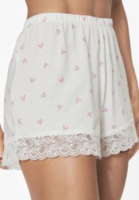 Weiße Baumwollshorts mit einem lila Herzmuster und einem gewellten Spitzenabschluss am Saum. Elastischer Taillenbund für zusätzlichen Komfort.