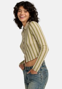 Joven mujer sonriendo, vistiendo un cárdigan corto de botones rayado beige y jeans de tiro alto azules, con las manos en los bolsillos, frente a un fondo sencillo.