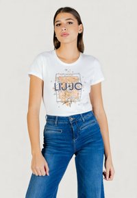 Camiseta blanca de algodón con estampado gráfico floral en colores naranja y azul, cuello redondo y mangas cortas, combinada con vaqueros de tiro alto en azul oscuro.