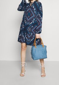 Blå mönstrad handväska med tanfärgade läderhandtag; intrikat blommönster i präglat mönster; buren med en marinblå mönstrad klänning och vita sandaler med remmar.