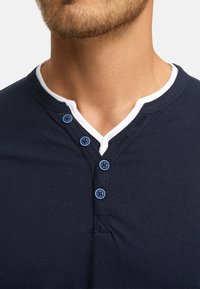Navy blue cotton henley shirt featuring a white collar and four blue buttons. The fabric is smooth with a fitted design.
