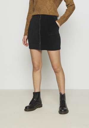 Kobieta w brązowym dzianinowym swetrze, czarnej mini spódnicy ze sztruksu z suwakiem z przodu, czarnych sznurowanych botkach, stoi na tle gładkiej ściany.