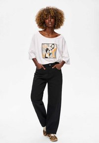 Biała, oversize'owa koszulka z grafiką psa w kwadratowym wzorze. Czarne szerokie spodnie. Buty sportowe w wzór lamparta.