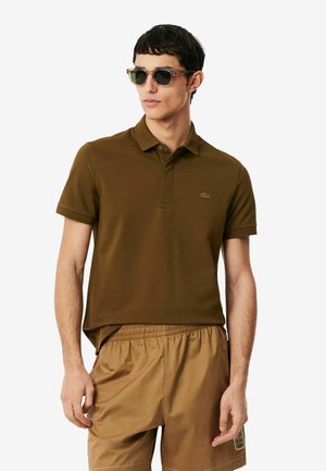 Joven con camisa tipo polo marrón, pantalones cortos color beige y gafas de sol, de pie frente a un fondo blanco liso.
