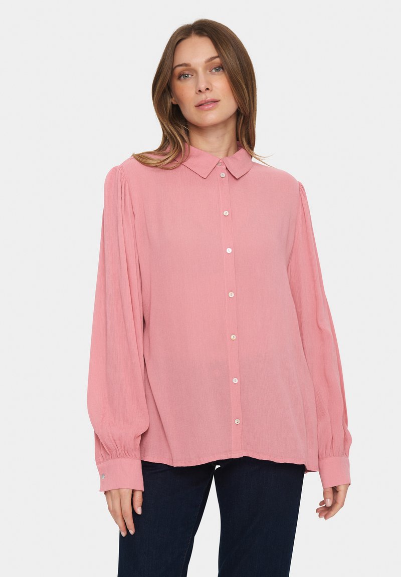 Camicia button-up a maniche lunghe di colore rosa chiaro con colletto, dettagli arricciati alle maniche e una texture morbida, abbinata a jeans in denim blu scuro.