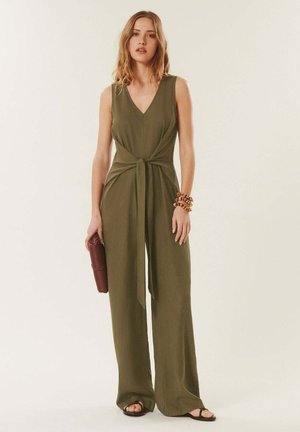 Femme en combinaison sans manches vert olive avec un détail noué à l'avant, tenant une pochette marron texturée, portant des bracelets et des sandales noires.