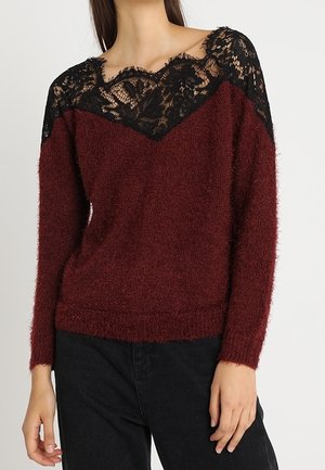 Femme portant un pull duveteux bordeaux avec un détail en dentelle noire sur la partie supérieure de la poitrine et les épaules, associé à un pantalon noir.