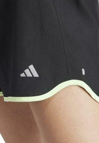 Pantalones cortos deportivos negros con un dobladillo ondulado, que presentan un ribete verde claro y un logo blanco cerca del lado inferior izquierdo. Tela suave y texturizada.