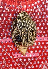 Czerwony materiał w tle ozdobiony iryzującymi koralikami. Centralna broszka w odcieniach złota z motywem ptaka, z czarnymi akcentami i skomplikowanymi detalami.