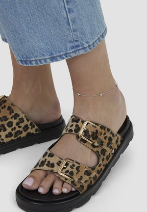 Füße in offenen Sandalen mit Leopardenmuster, goldenen Schnallen und einer silbernen Herzknöchelketten, kombiniert mit hellblauen Jeanshosen.